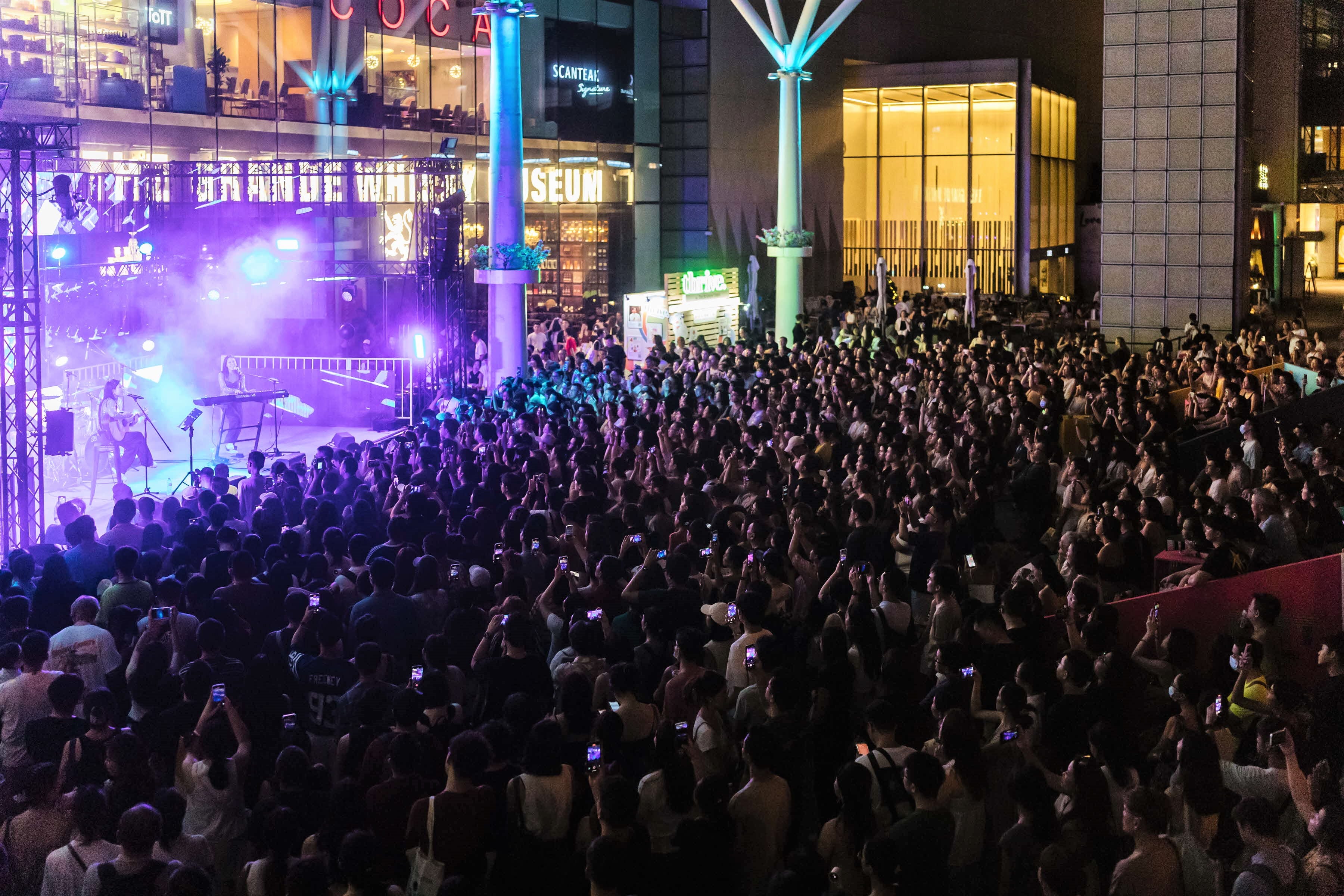 Nighttime crowd at outdoor concert. Stage lights illuminate performers, audience holding up phones. Modern buildings in background.